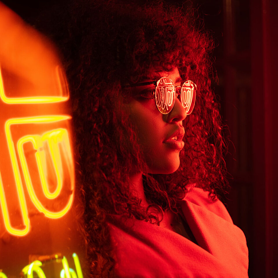 woman with large curly hair standing beside a neon sign with sunglasses on