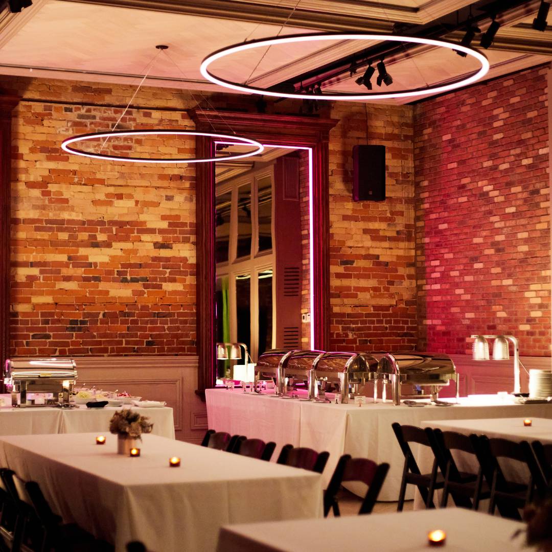 Elegant event venue with exposed brick walls, banquet tables dressed in white linens, buffet stations, and modern circular pendant lighting creating a warm ambient glow.