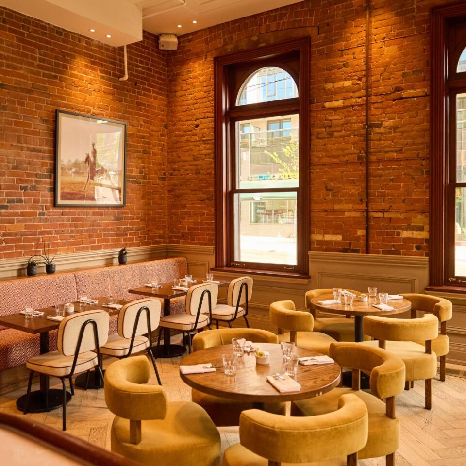 Warm and inviting restaurant dining room with exposed brick walls, large arched windows, round wooden tables, and plush mustard-coloured chairs in a historic setting.