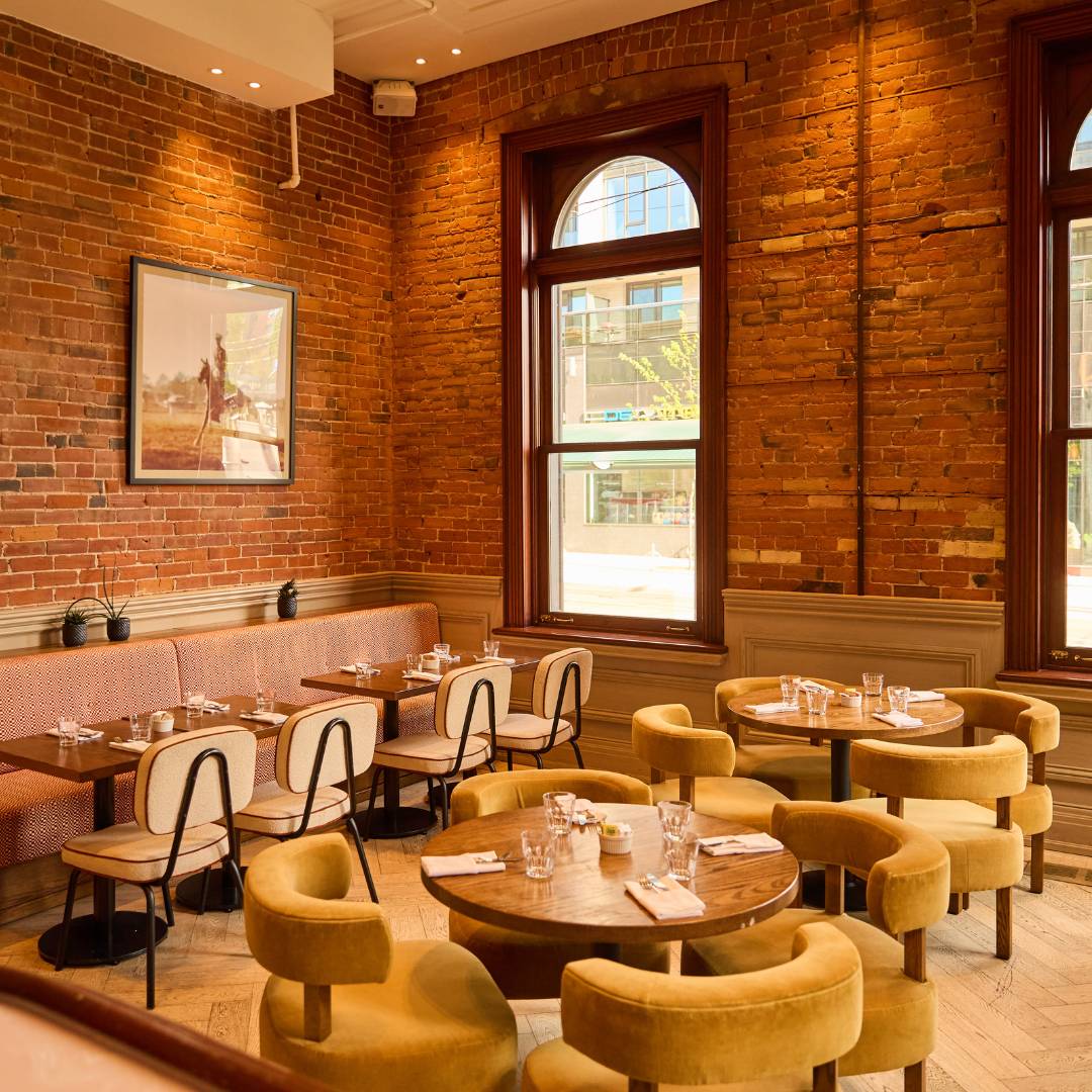 Warm and inviting restaurant dining room with exposed brick walls, large arched windows, round wooden tables, and plush mustard-coloured chairs in a historic setting.
