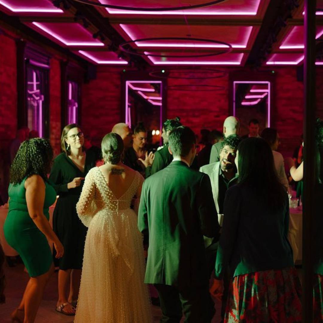 Guests socializing at an evening event in a historic venue, illuminated by vibrant pink accent lighting and modern ceiling fixtures, creating an energetic and elegant atmosphere.