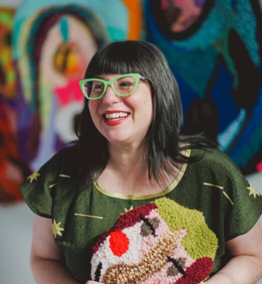 Smiling woman with black hair and green glasses holding a colourful textile artwork in front of vibrant abstract wall art.