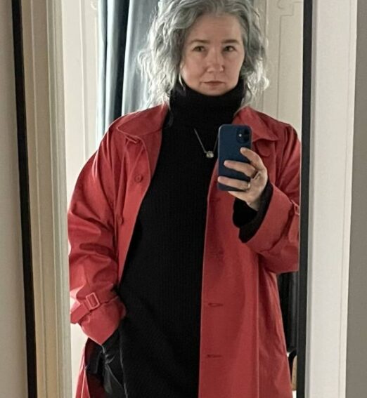 Woman with wavy grey hair taking a mirror selfie while wearing a red coat over a black outfit.