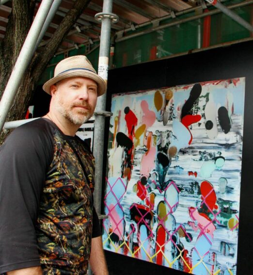 Man wearing a fedora standing beside a colourful abstract painting displayed outdoors.