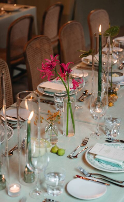 Elegant dining table set with green taper candles, colourful flowers, glassware, and patterned place settings.