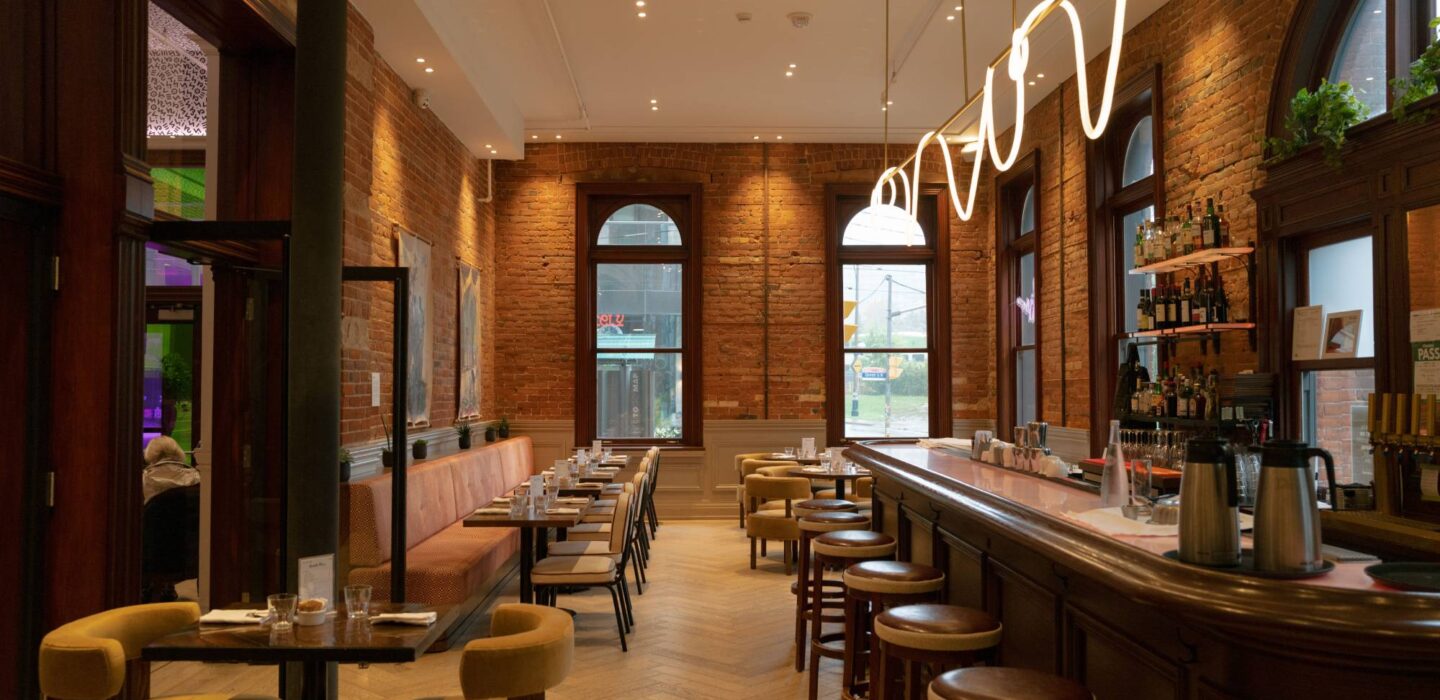 Modern restaurant interior with exposed brick walls, a long wooden bar, upholstered seating, and sculptural pendant lighting.