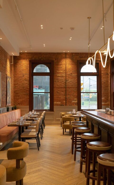 Modern restaurant interior with exposed brick walls, a long wooden bar, upholstered seating, and sculptural pendant lighting.