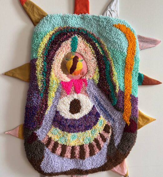 Colourful tufted textile wall hanging depicting a stylized figure with an eye motif, surrounded by fabric spikes.