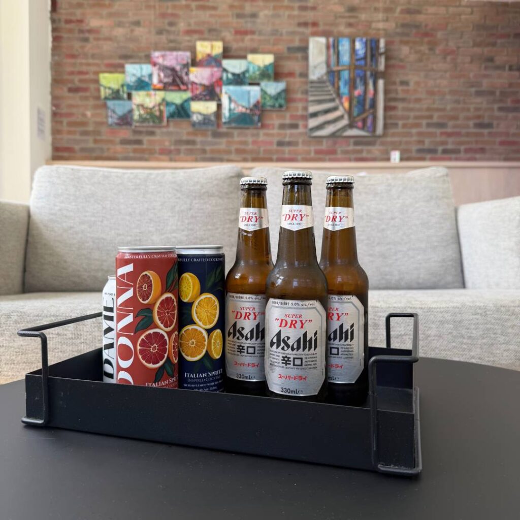 Bottles of Asahi Super Dry beer and colourful Italian spritz cans arranged in a black tray on a table, with lounge seating and artwork in the background.
