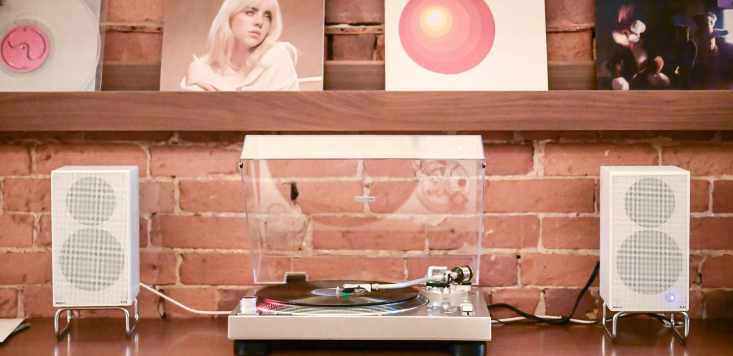 Turntable playing a vinyl record between two white speakers, with album covers displayed on wooden shelves against an exposed brick wall.