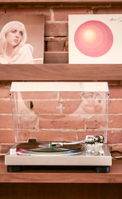 Turntable playing a vinyl record between two white speakers, with album covers displayed on wooden shelves against an exposed brick wall.