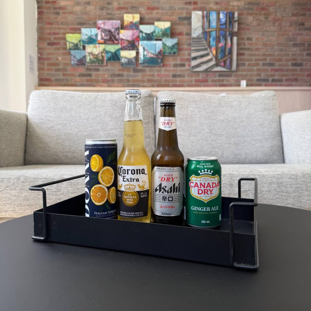 A Corona Extra bottle, Asahi Super Dry beer, Canada Dry ginger ale, and an Italian spritz can arranged in a black tray on a table, with lounge seating and colourful artwork behind them.
