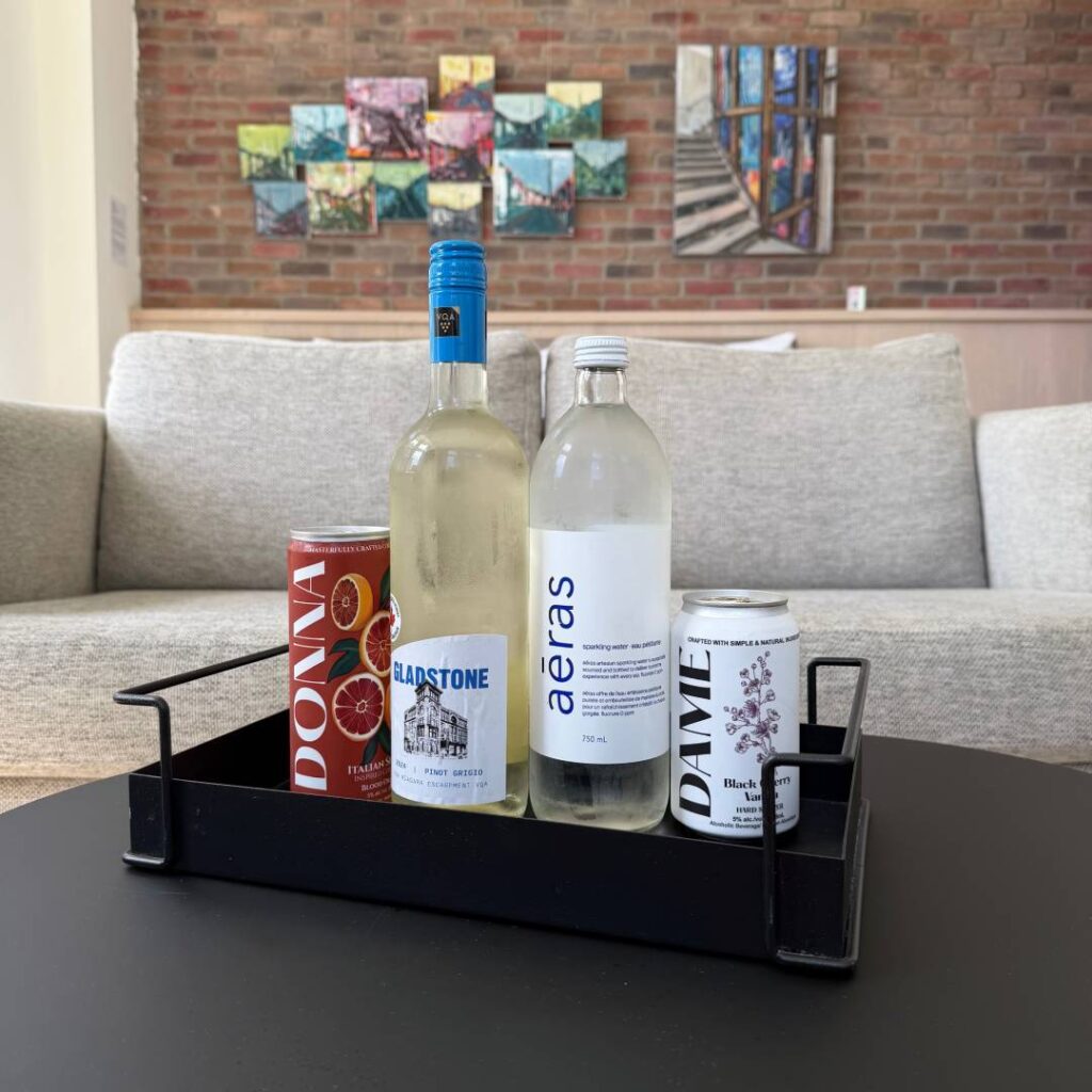 Bottles of Gladstone Pinot Grigio and aēras sparkling water with two colourful canned cocktails arranged in a black tray on a table, with lounge seating and artwork in the background.