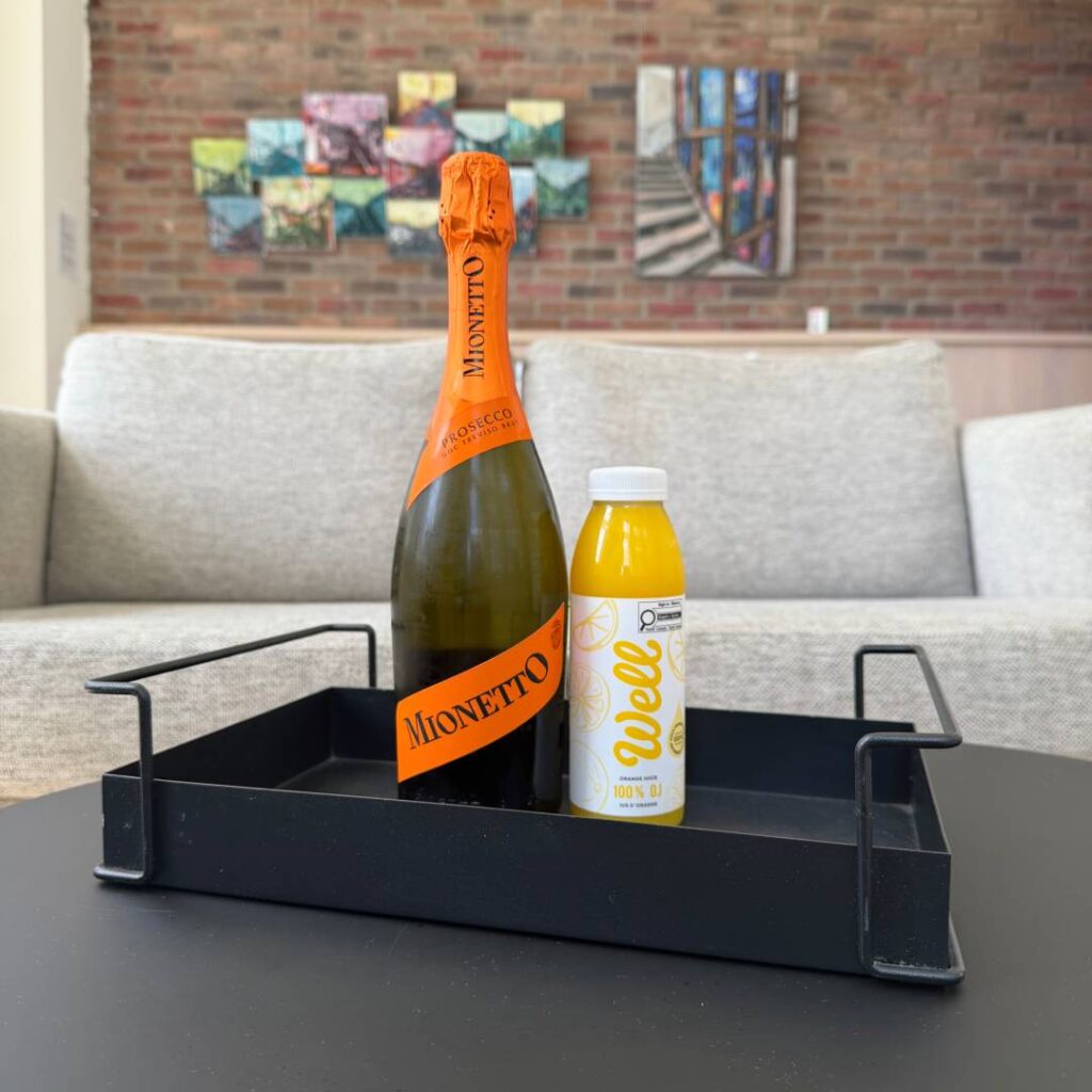 Bottle of Mionetto Prosecco and a small bottle of orange juice arranged in a black tray on a table, with lounge seating and colourful artwork in the background.