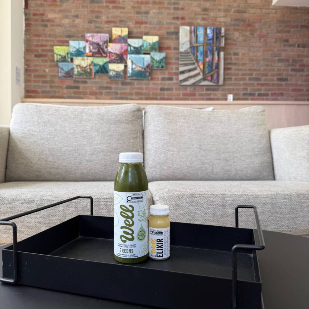 Bottle of Well Greens juice and a small Well Elixir wellness shot arranged in a black tray on a table, with lounge seating and colourful artwork in the background.